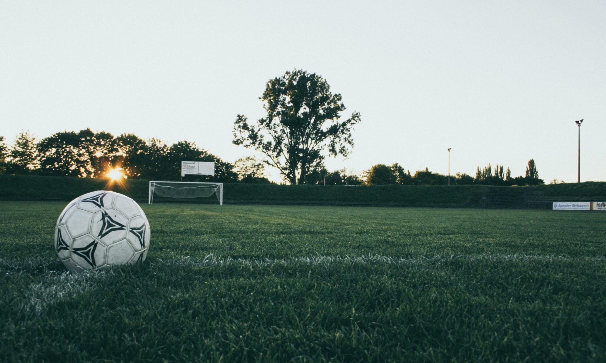 Balón de fútbol sobre el césped de un campo al atardecer, con la portería al fondo.